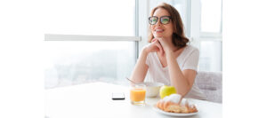 Picture image of cheerful young pretty woman sitting indoors at the table with juice and croissant eating corn flakes. Looking aside.